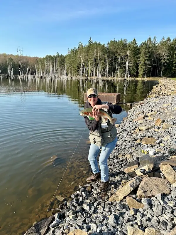 Amy smiling while holding a large trout on a sunny lake, representing a perfect day and choosing a better life over constant work